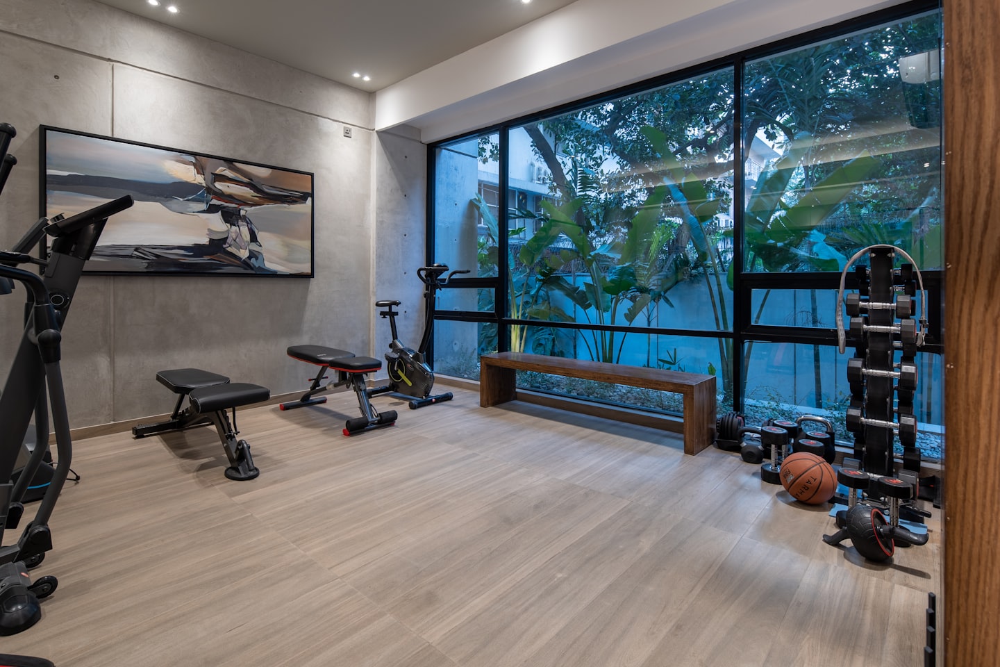 A home gym with exercise equipment, a wooden bench, and large windows overlooking plants