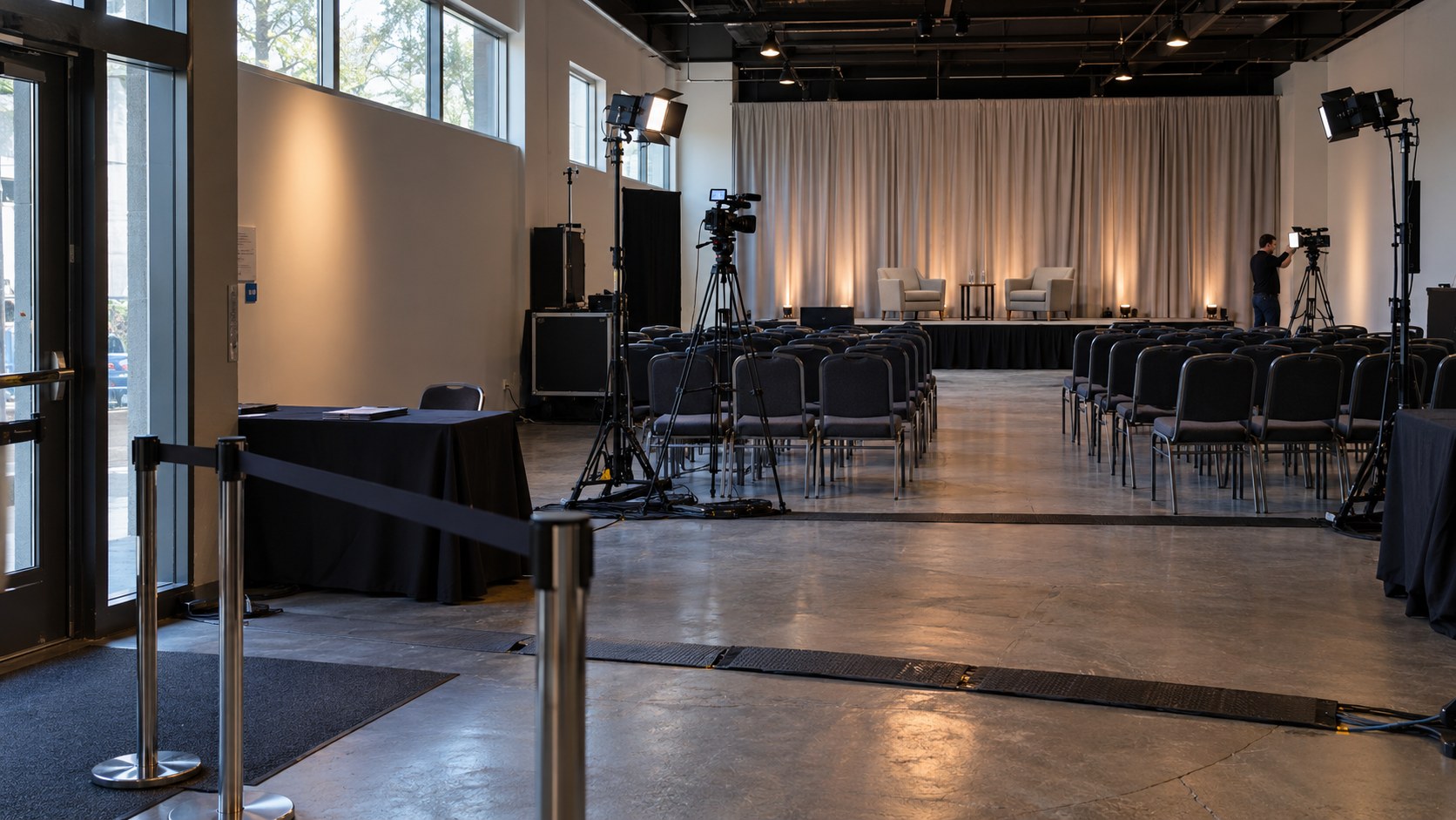 Wide view of a compact broadcast venue with an entry checkpoint, audience chairs, cameras, stage lights, cable ramps, and a technician testing equipment.