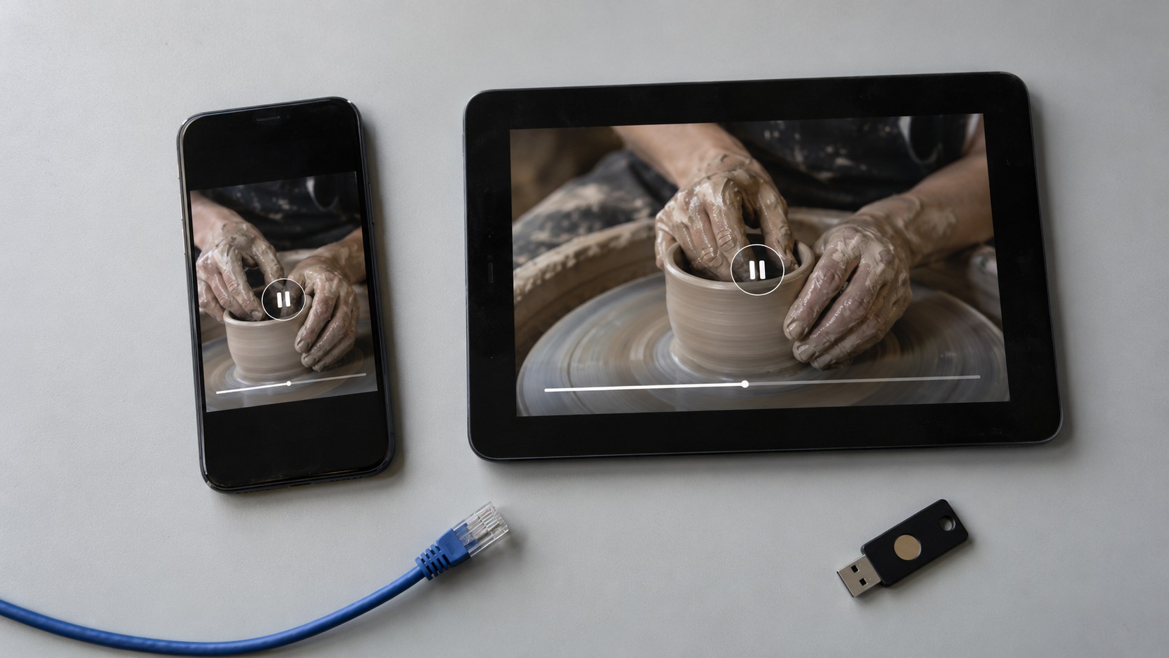 Overhead view of a phone and tablet showing the same paused pottery lesson beside a disconnected network cable and USB security key.